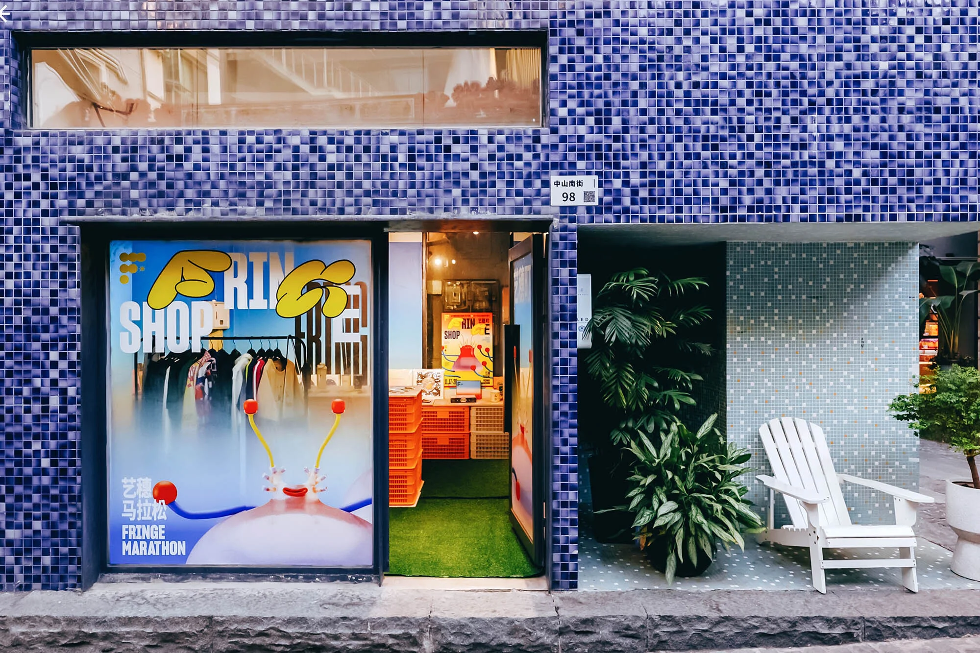 Exterior of Fringe Marathon store with blue tiled facade, open door, display window, plants, and white chair on a paved walkway.