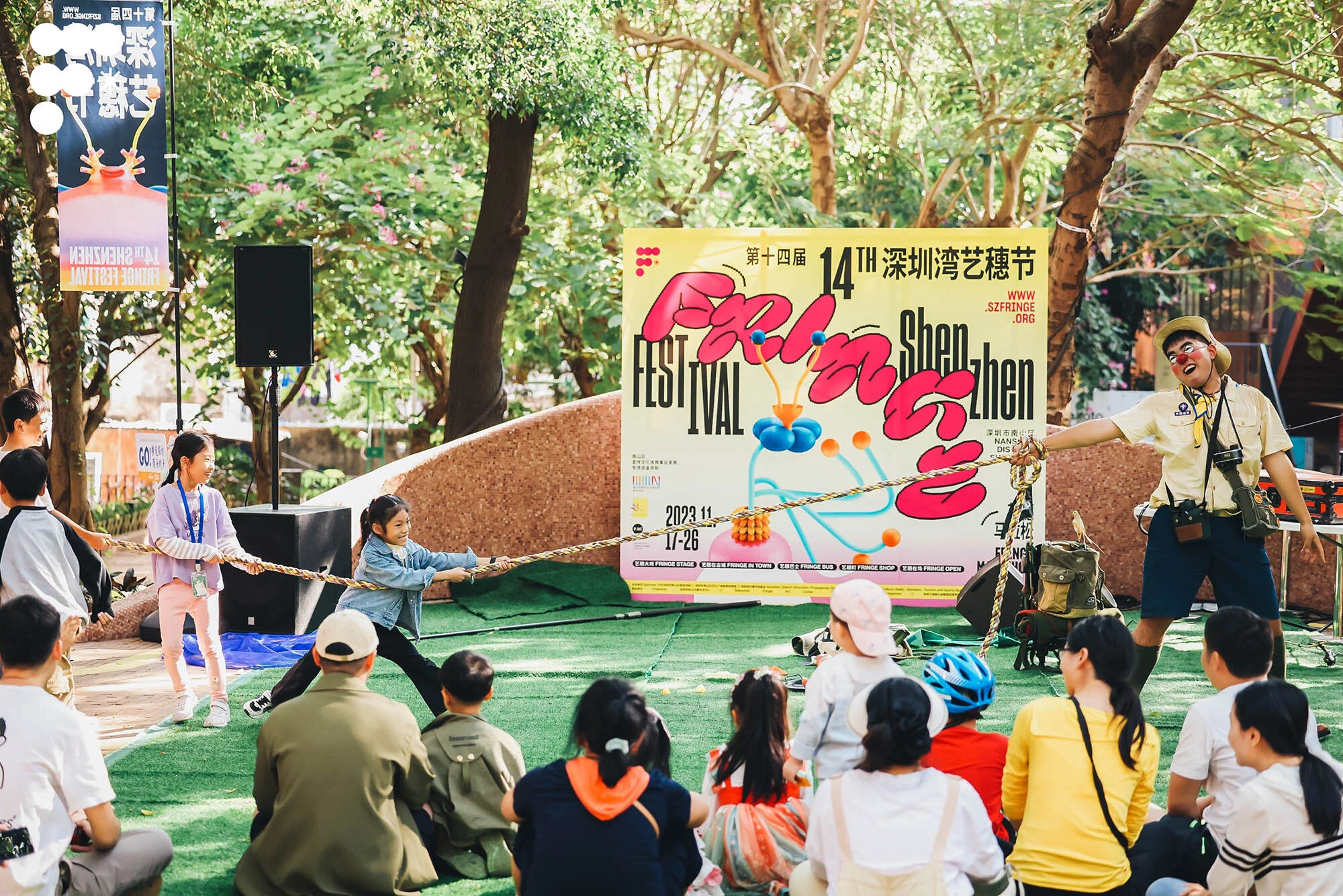Outdoor festival scene with children and a clown engaging in a tug-of-war on artificial grass, against a backdrop of trees and event signage.