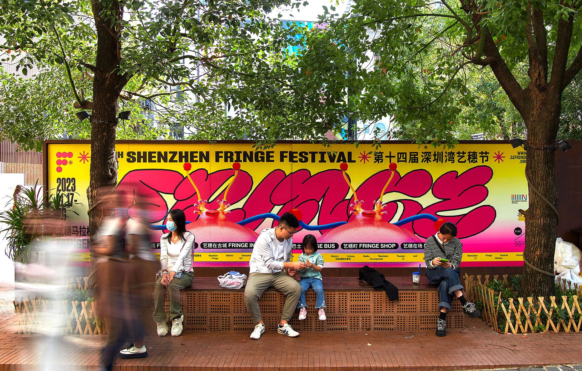 Outdoor shot of people sitting on a bench in front of a Shenzhen Fringe Festival banner, blurred pedestrians passing by, lush green trees.