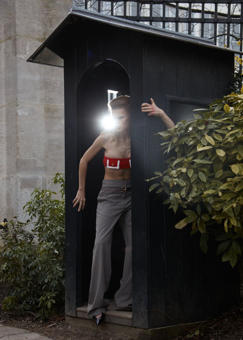 A woman in a red tube top and grey pants stands in a dark-painted wooden telephone booth surrounded by greenery and stonework.