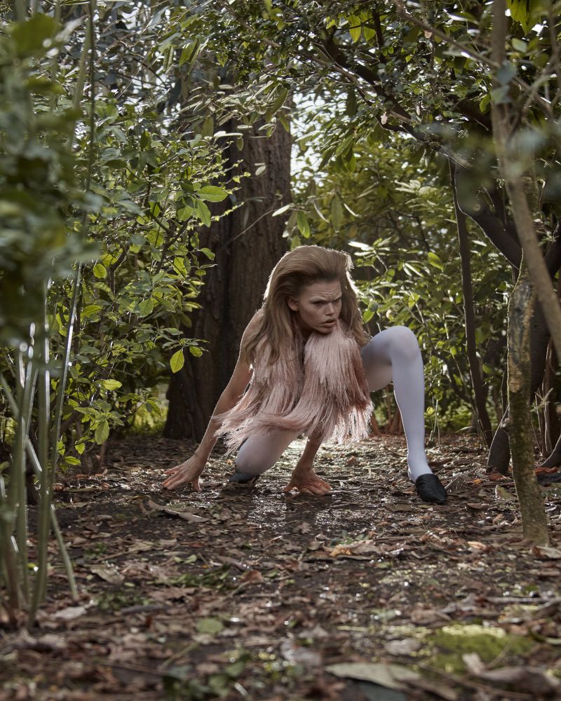 An adult female crouches in a lush forest setting wearing a fringe top, white tights, and black shoes for a fashion editorial shot.