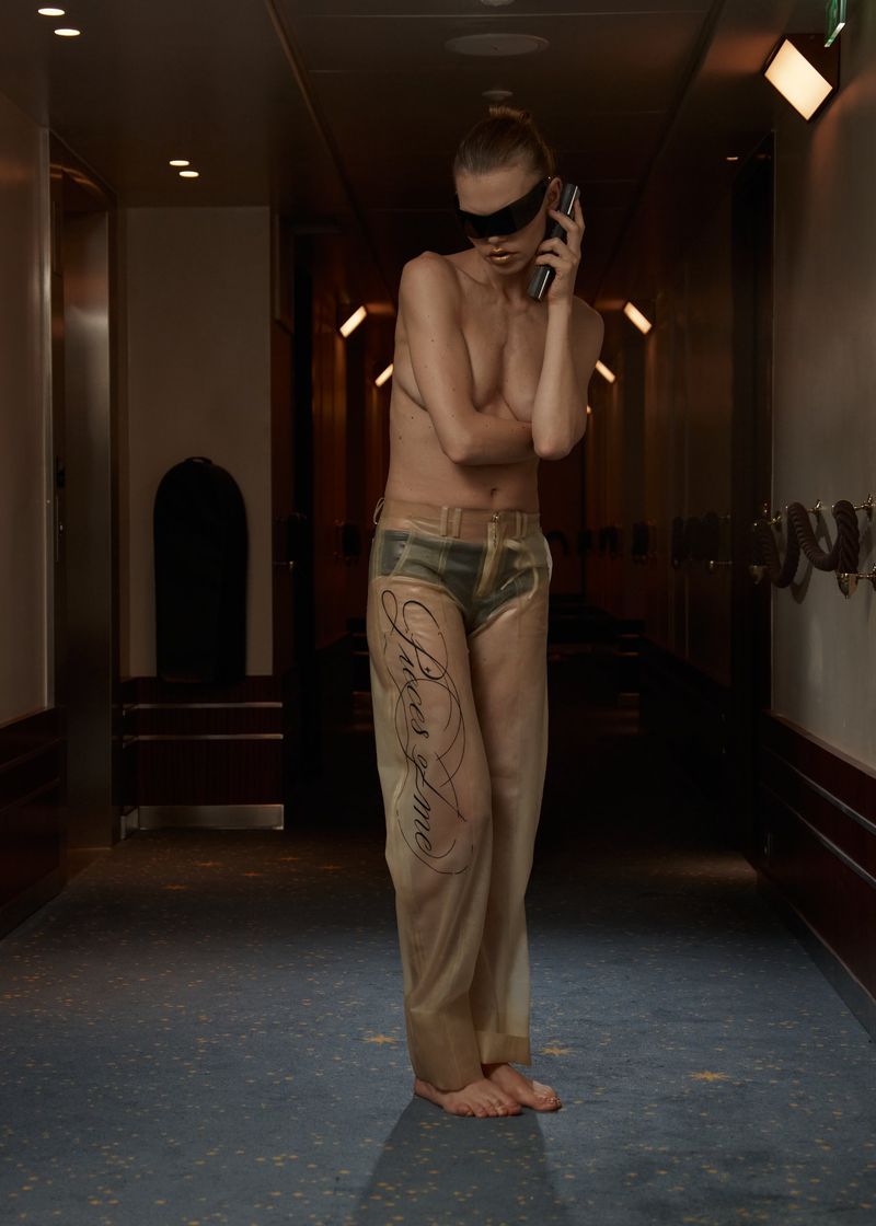 Blonde woman with blindfold and body paint in a hallway holding a phone. The artwork features tattoo-style letters and camouflage patterns.