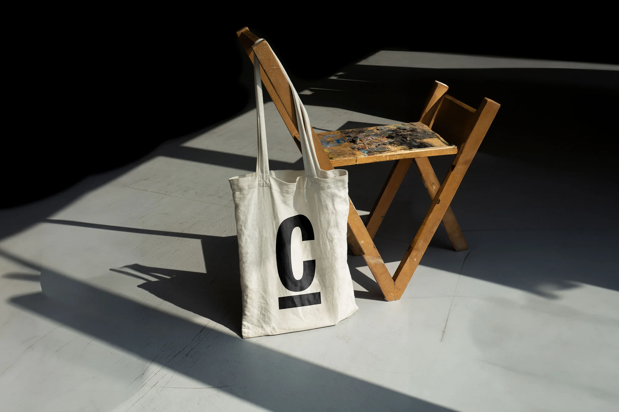 Canvas tote bag with a large 'C' hangs on a folding chair in a photo studio setting, featuring strong shadows and studio lighting.