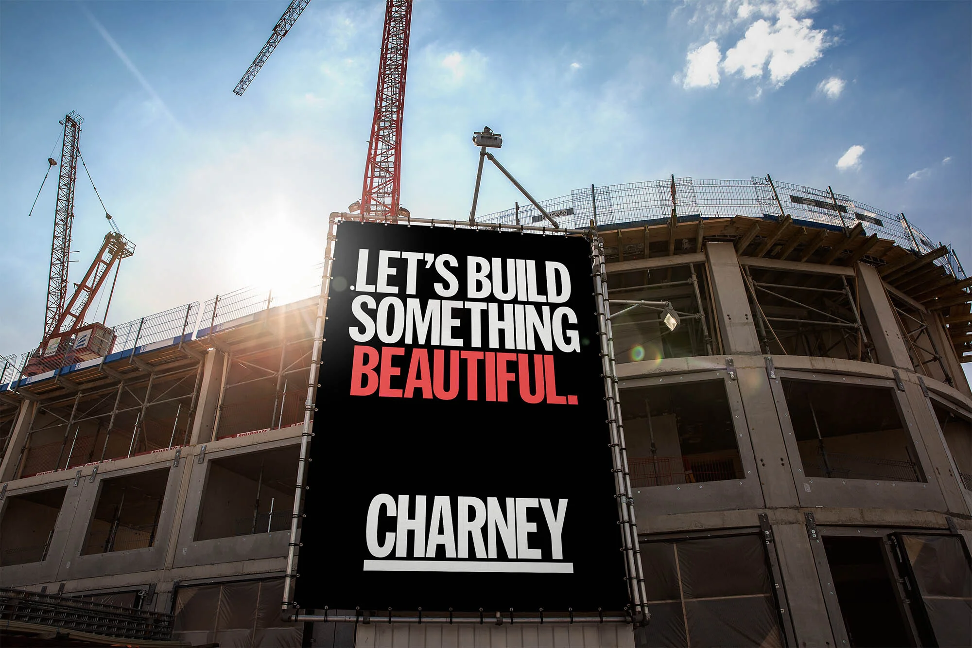 Construction site with a large black billboard stating, 'Let's Build Something Beautiful. Charney.' with red cranes and blue sky background.