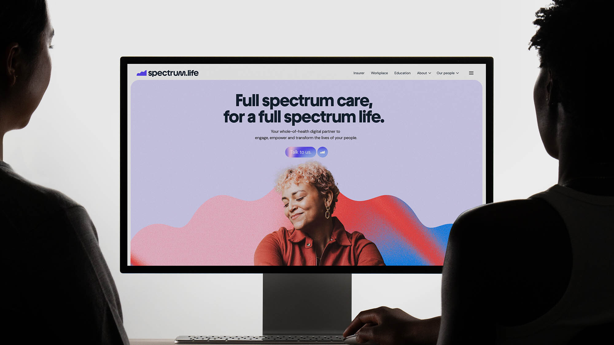 Two people looking at a computer monitor displaying the Spectrum.life website with a woman and colorful graphics.