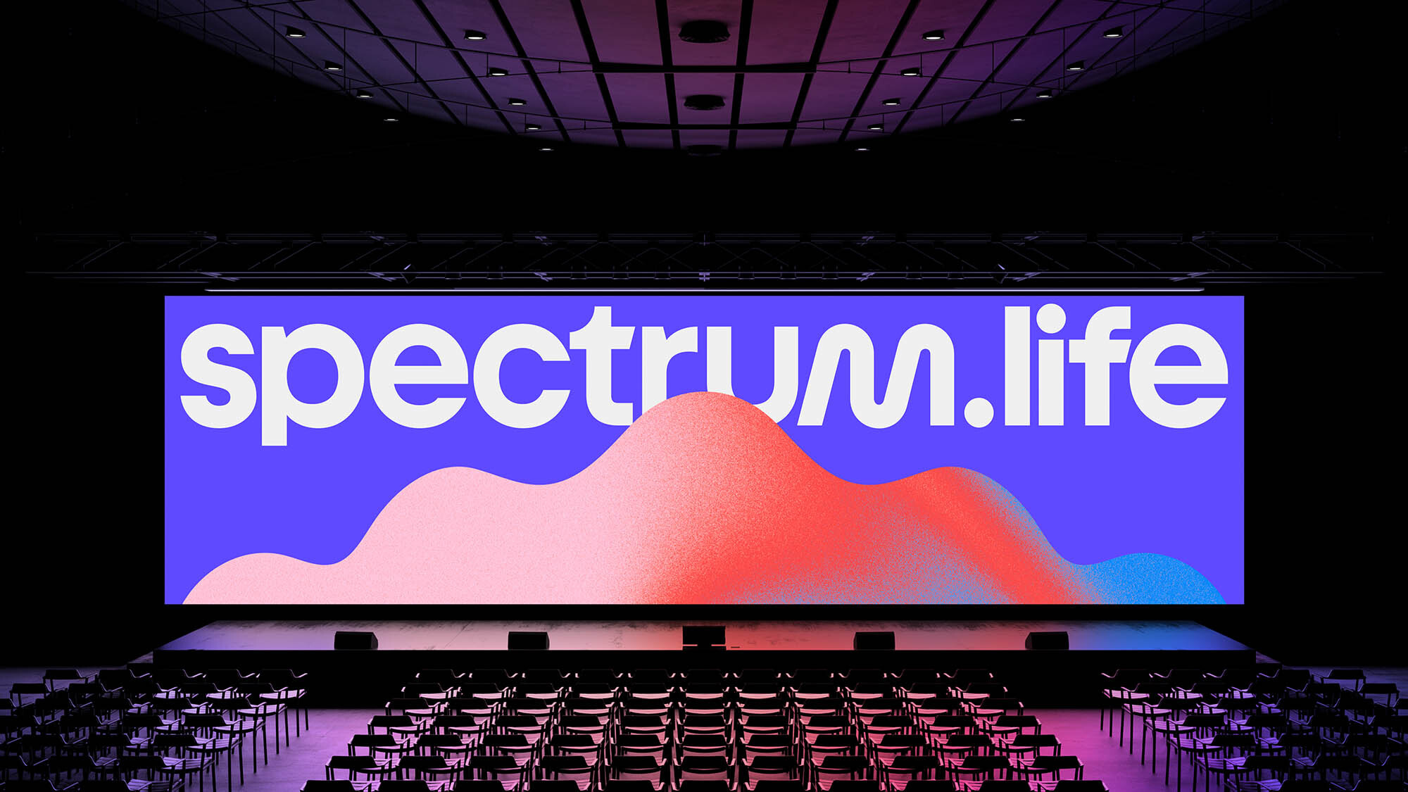 Empty auditorium with rows of chairs facing a large screen displaying 'spectrum.life' logo and a wave graphic, lit with stage lighting.