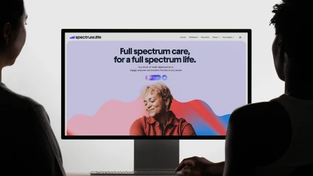Two people viewed from behind looking at a computer monitor displaying the Spectrum Life website homepage