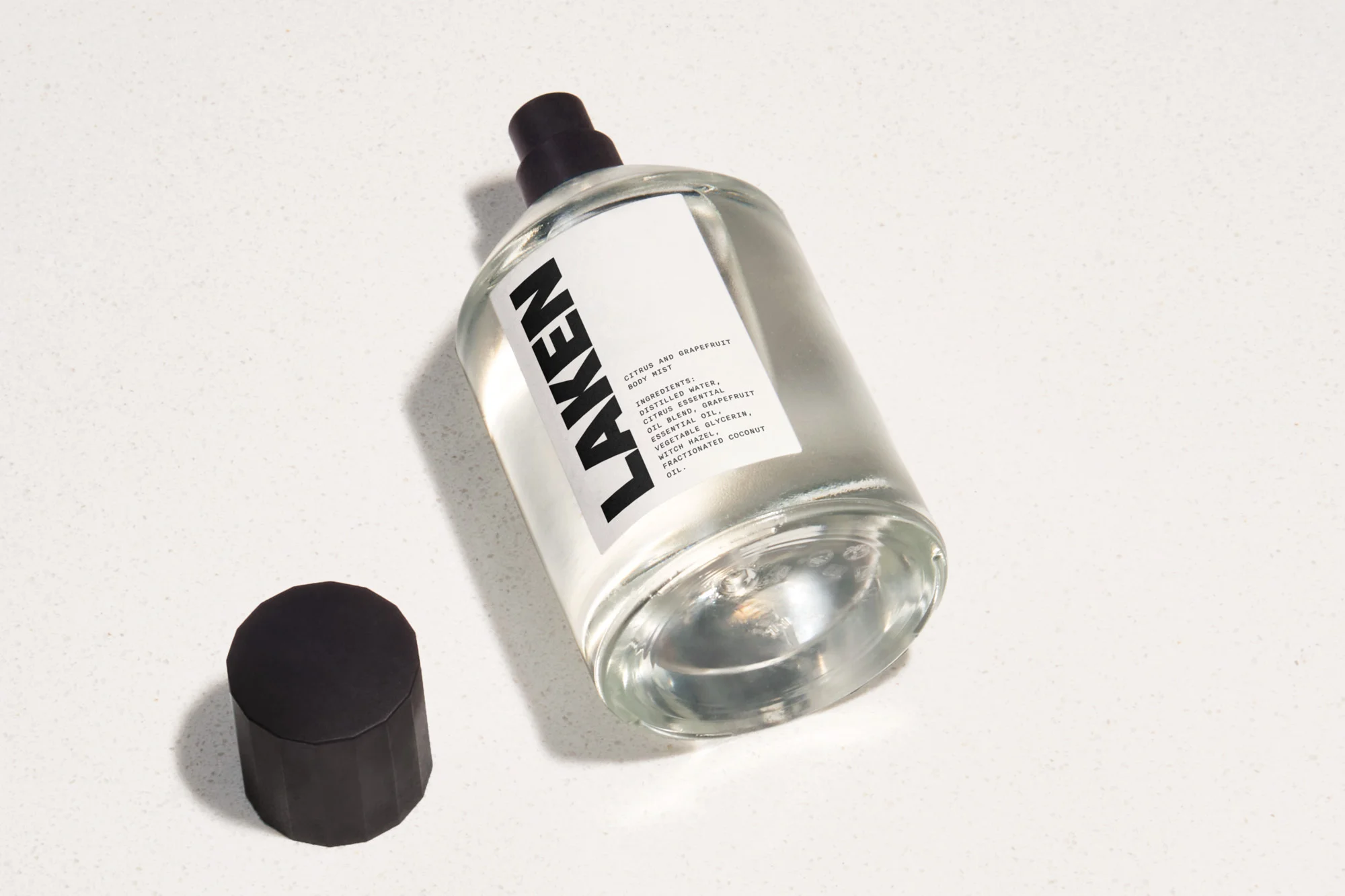 Clear glass Laken spray bottle with black cap sits beside its black cylindrical cap on a clean white surface.