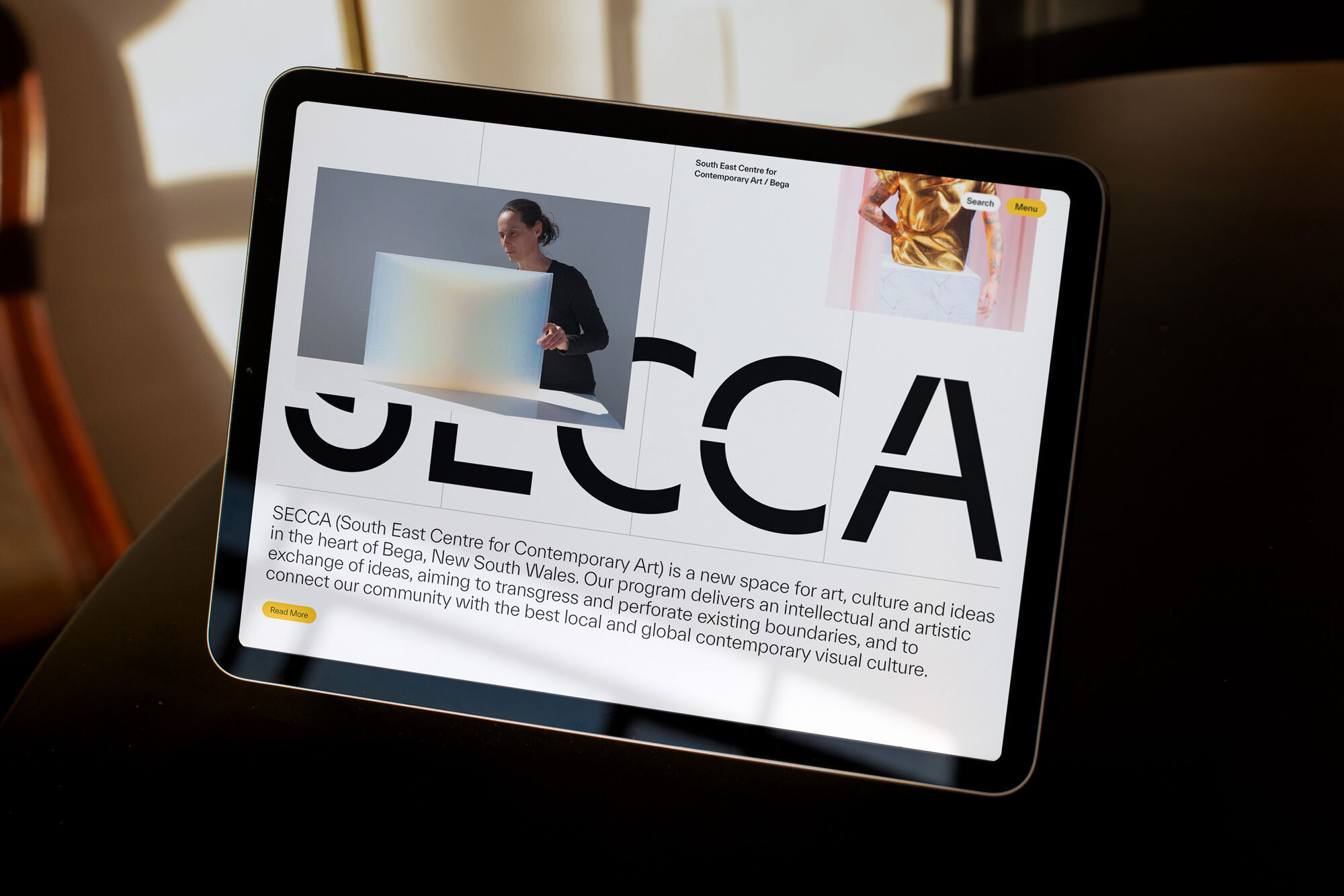 An iPad displays the SECCA (South East Centre for Contemporary Art) website featuring an artwork image and site navigation elements.