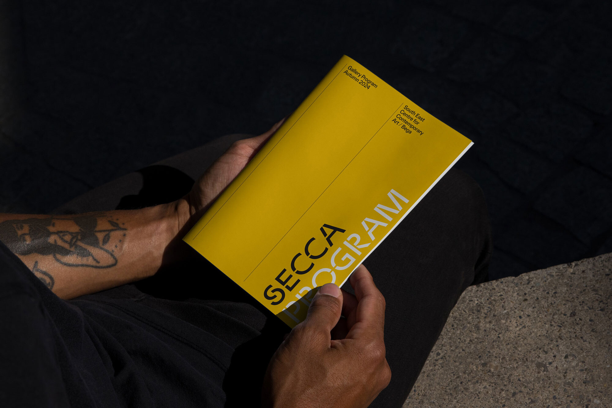 Adult hands hold a SECCA Program booklet with a yellow cover, sitting outdoors on a stone surface with dark shadow.