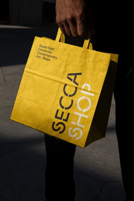 Close-up of a yellow paper shopping bag with the words 'SECCA Shop' on it, held by an adult in a dark business suit.