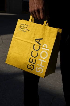 Close-up of a yellow paper shopping bag with the words 'SECCA Shop' on it, held by an adult in a dark business suit.