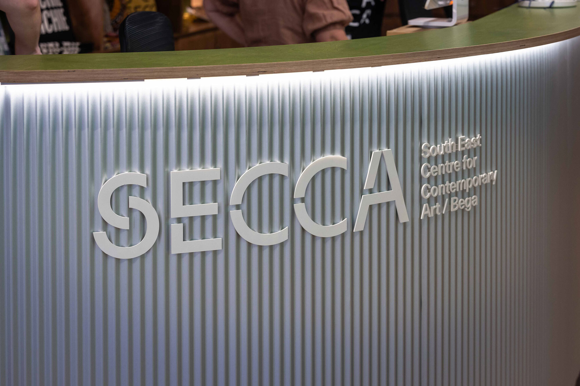 Interior view of SECCA's contemporary art center reception area with a corrugated wall, logo signage, and illuminated desk.