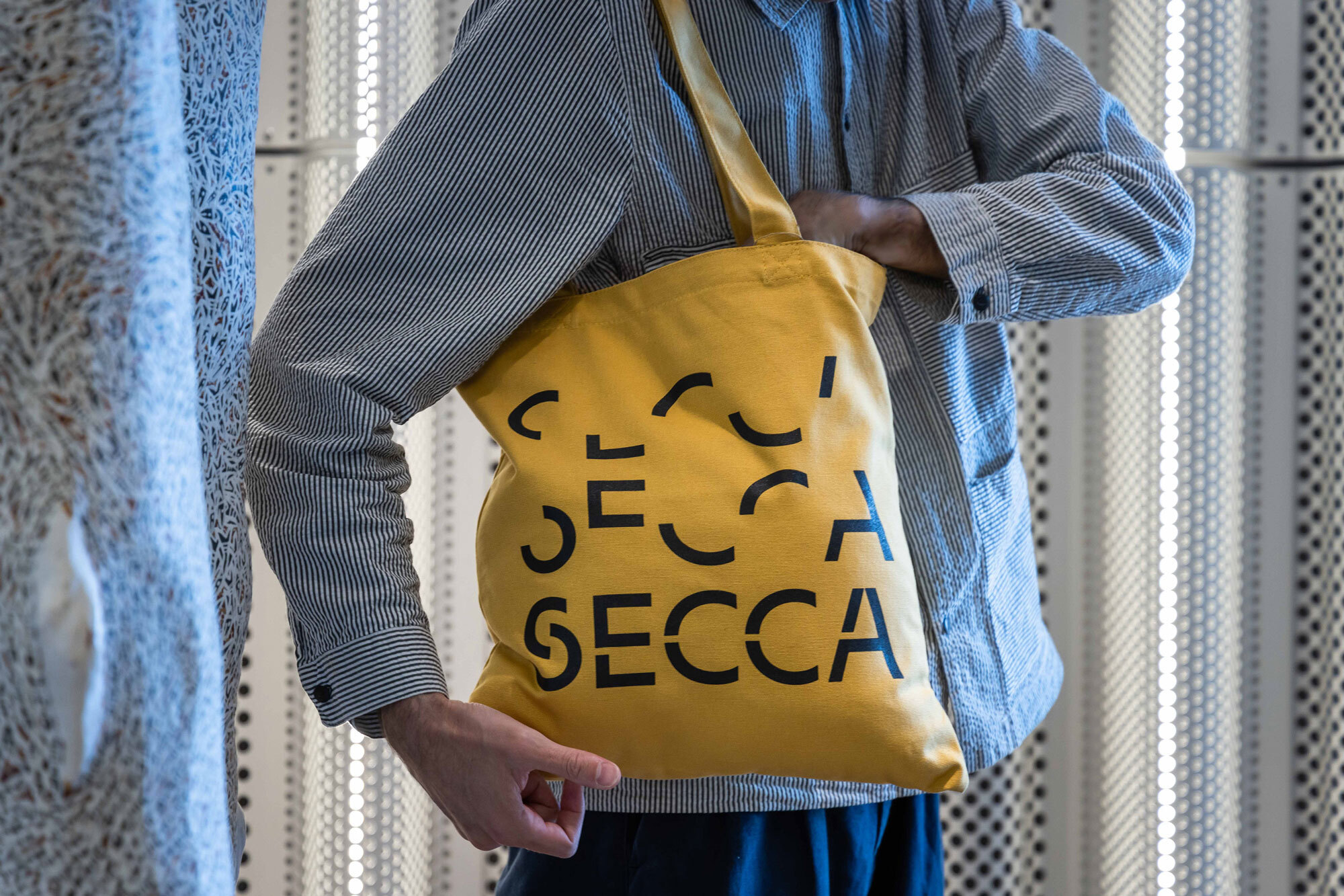 Person holds yellow SECCA tote bag in a striped shirt against a textured background, creating a casual and modern aesthetic.