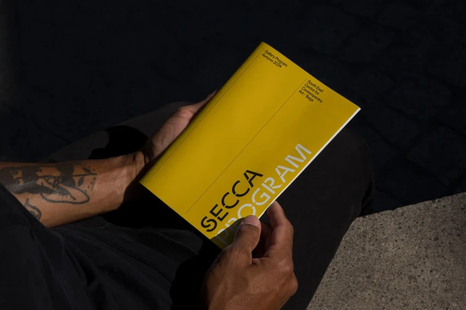 Person holding a bright yellow SECCA Program brochure outdoors in direct sunlight with dark clothing and visible arm tattoo.
