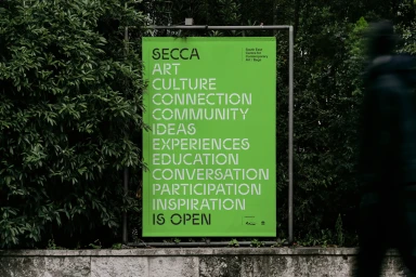 A vibrant green outdoor poster for SECCA displaying bold white typography amidst lush green foliage and a concrete wall.