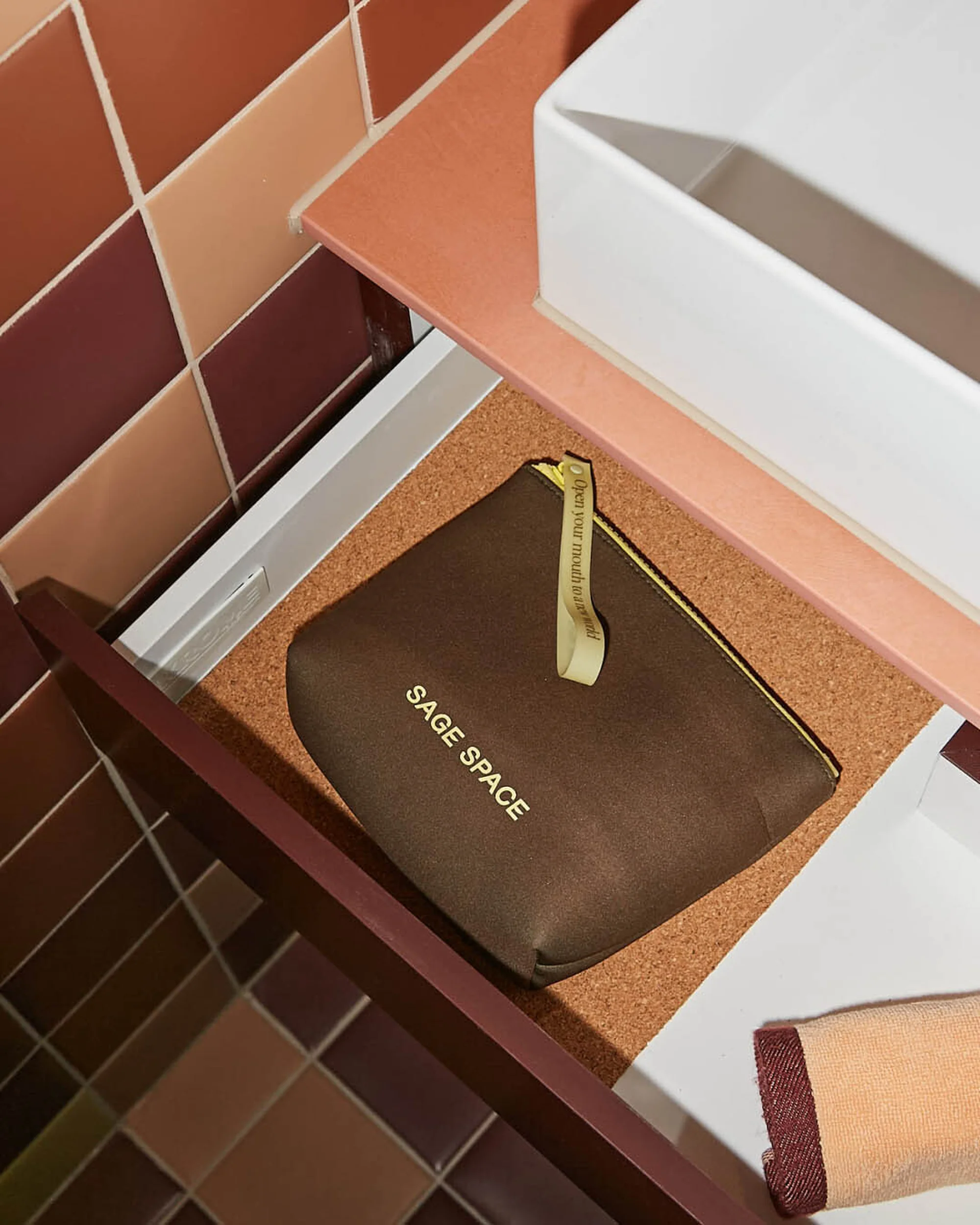 A sage green cosmetic bag sits inside an open drawer, next to a white sink and colorful tiled wall in a bathroom setting.
