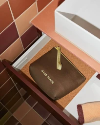 Brown cosmetic pouch with white Sage Space text resting inside an open drawer lined with cork, beneath a white sink