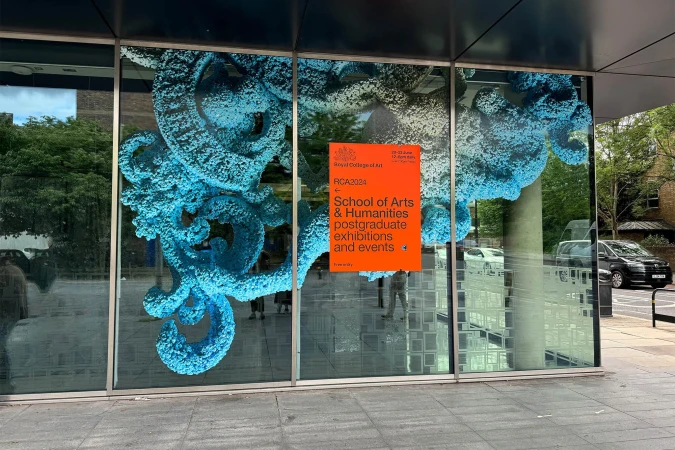 Building window display featuring abstract blue sculpture and orange Royal College of Art postgraduate exhibitions and events advertisement.