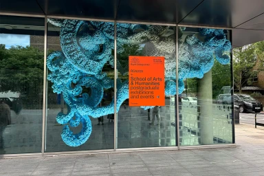 Large blue textured sculptural installation displayed behind glass windows of a building exterior with an orange event poster
