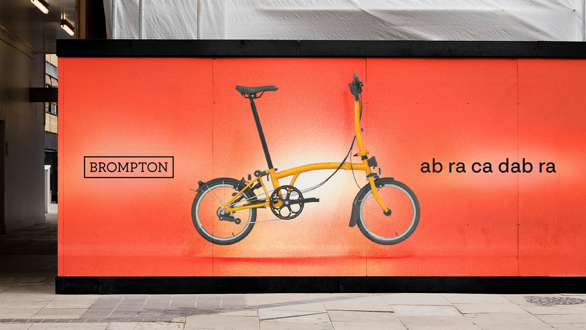 Yellow Brompton folding bicycle on orange background wall with Brompton logo and 'ab ra ca da bra' text, photographed outdoors.