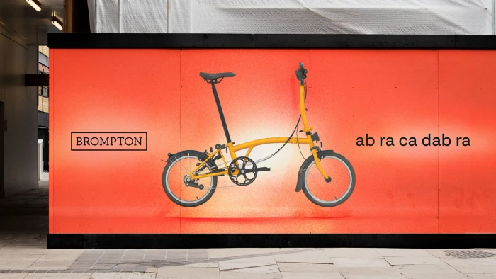 A yellow folding bicycle displayed on a bright orange promotional outdoor hoarding for Brompton Bicycles.