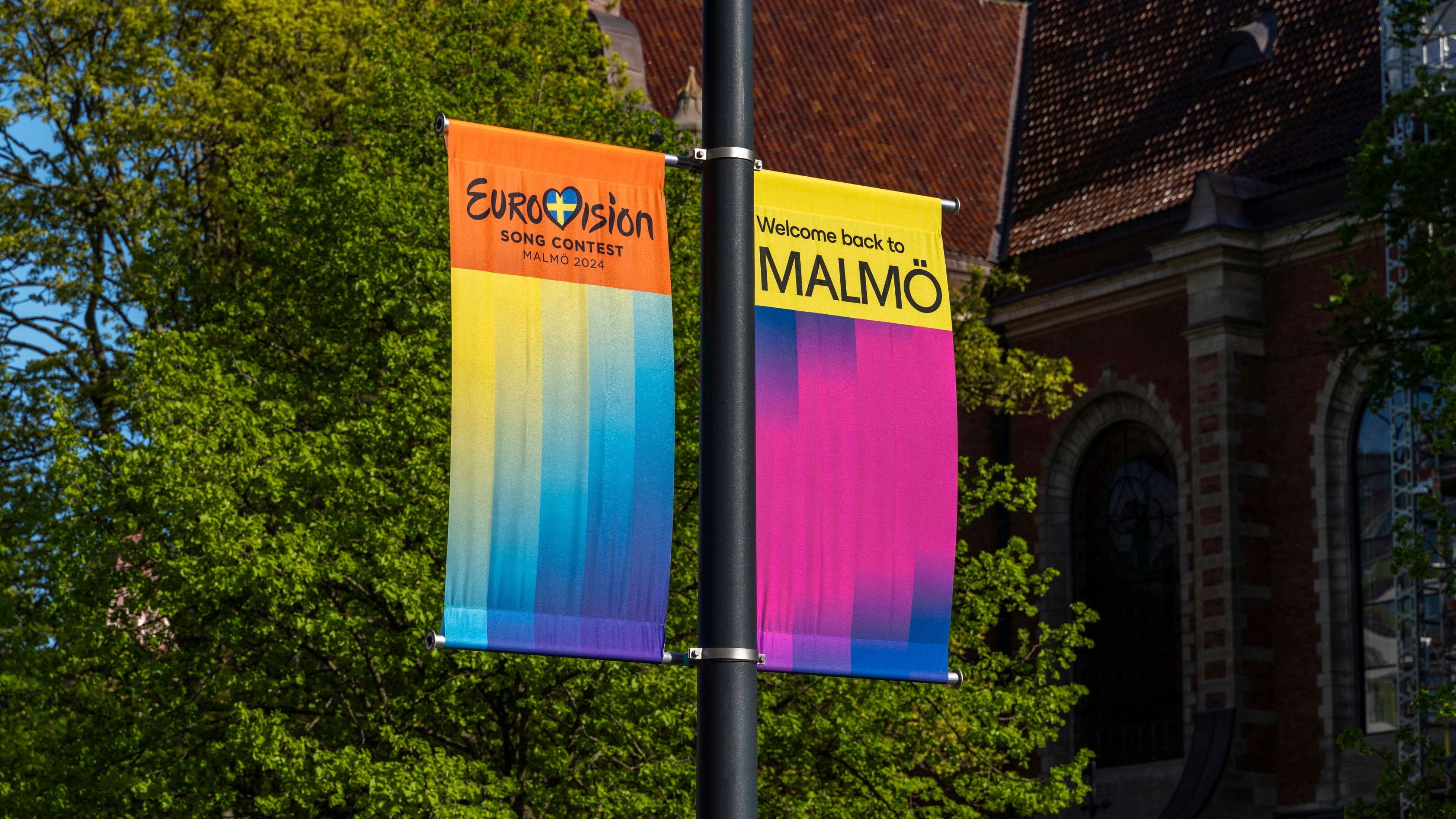 Two colorful Eurovision banners hang on a pole in Malmo, Sweden. Lush green trees and a brick building form the backdrop.