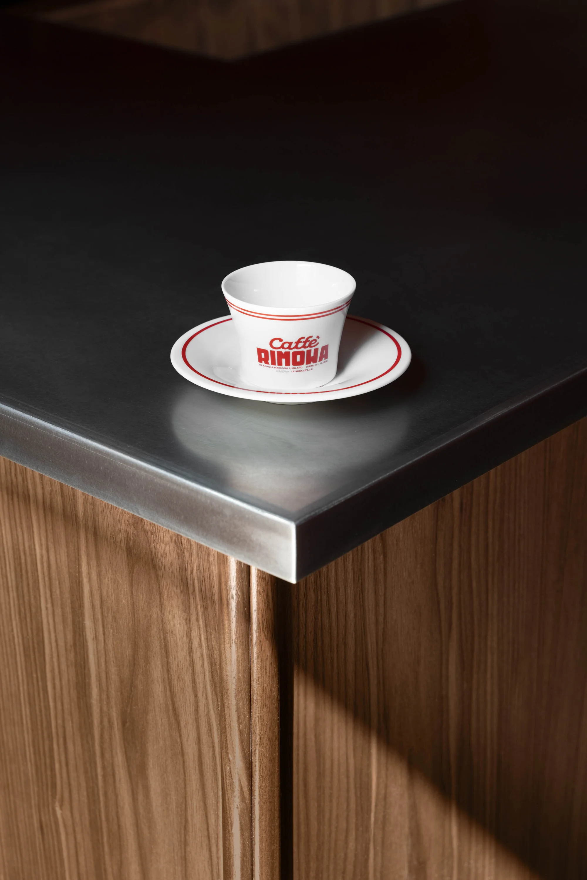 Close-up of a white coffee cup and saucer with red trim and 'Caffe RIMOVA' text, placed on a dark countertop with wood cabinetry.