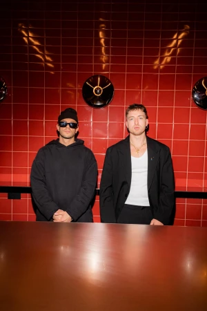 Two adult men pose indoors against a red tile wall; one wears sunglasses and a beanie while the other wears a blazer and necklace.