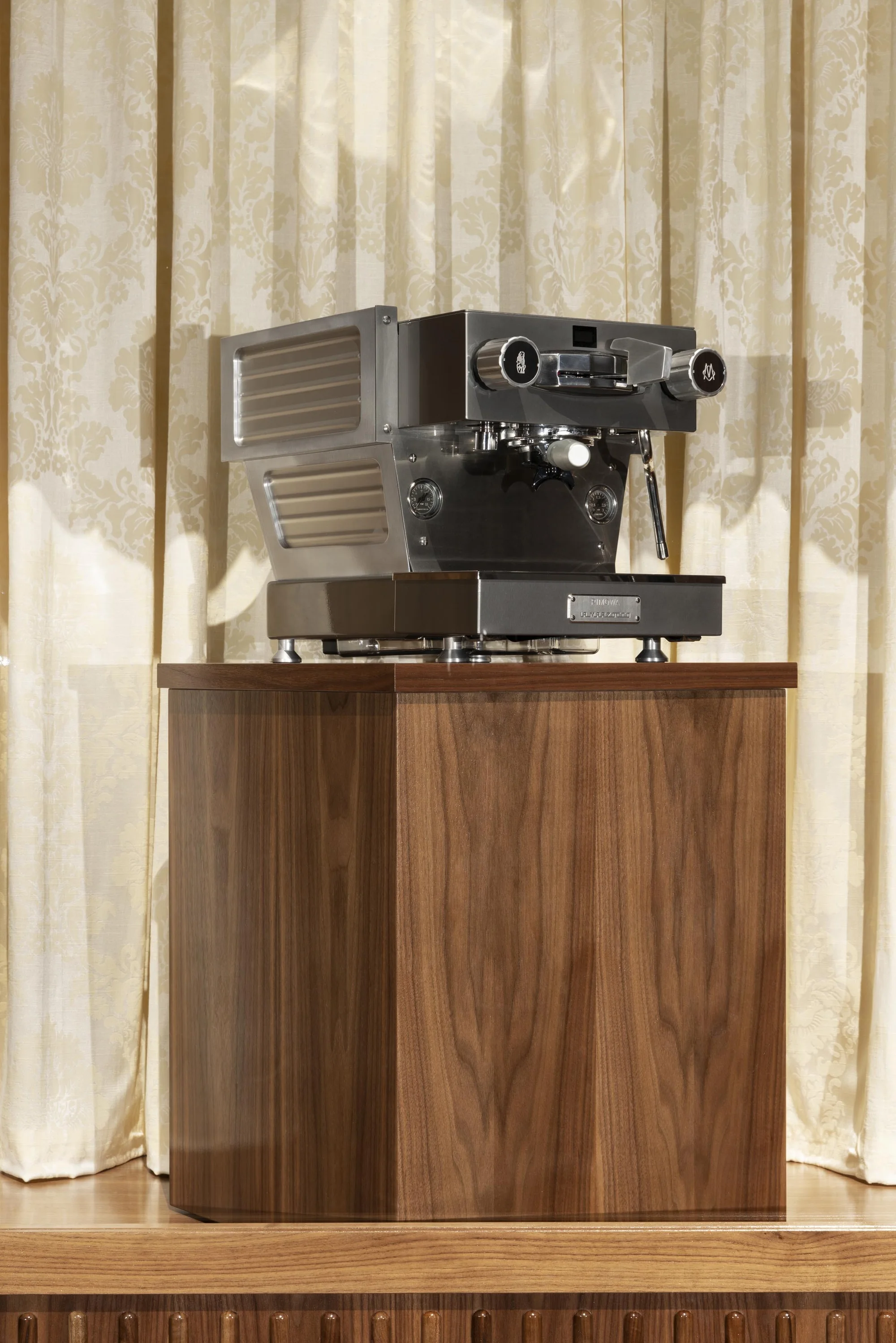 An espresso machine is displayed on top of a wooden cabinet with cream-colored curtains in the background, indoor setting.