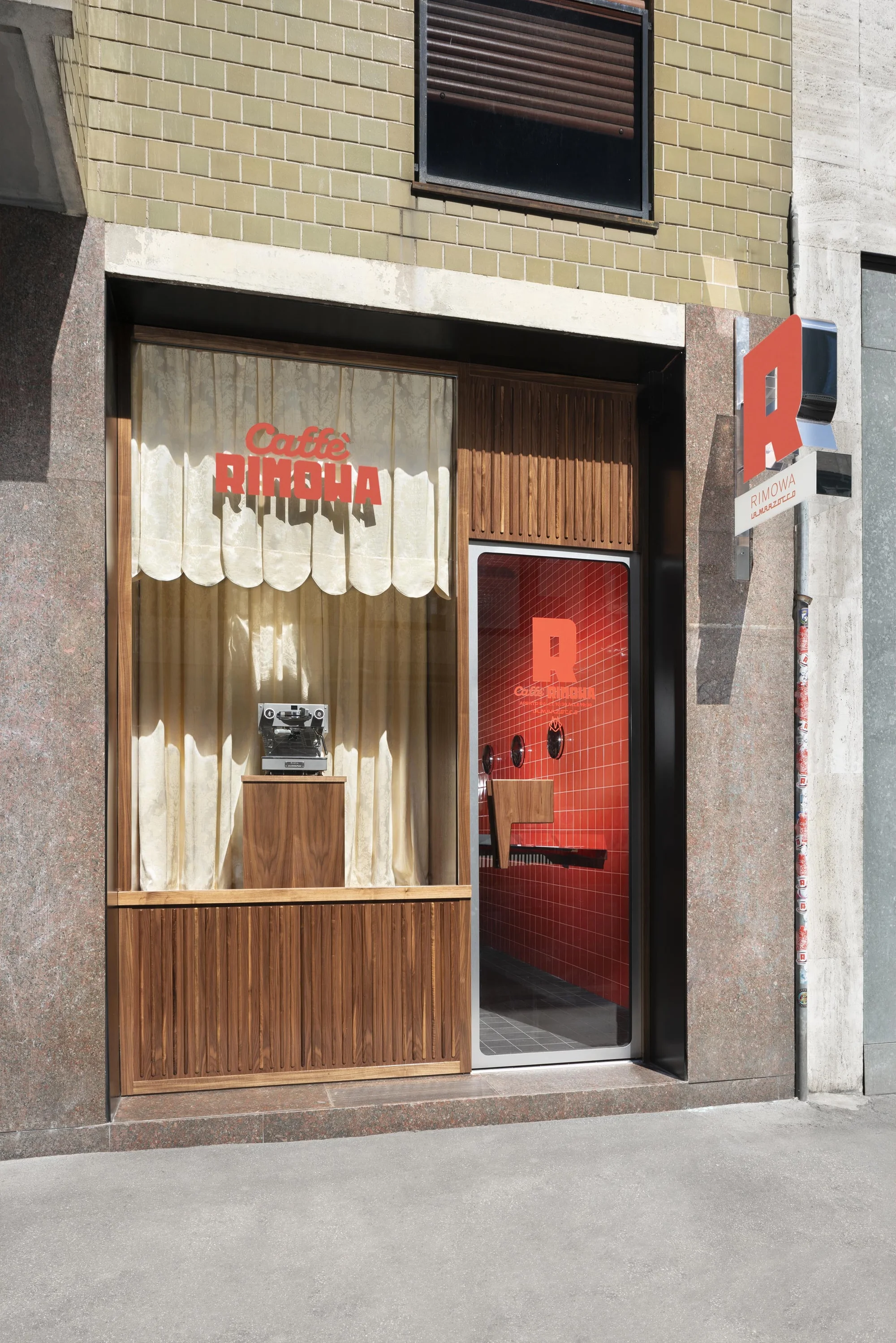 Exterior view of Caffe Rimowa featuring a window display with vintage television and cream-colored curtains in a modern urban setting.