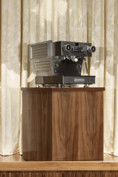 A metallic commercial espresso machine with ridged side panels resting on a wooden pedestal against patterned curtains.