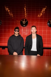 Two men standing side-by-side against a glossy red tiled wall, one wearing a black hoodie and beanie, the other in a black blazer.