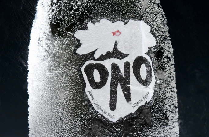 Close-up of a frosty sake bottle with a white label featuring a flower and the word "ONO," set against a dark, contrasting background.