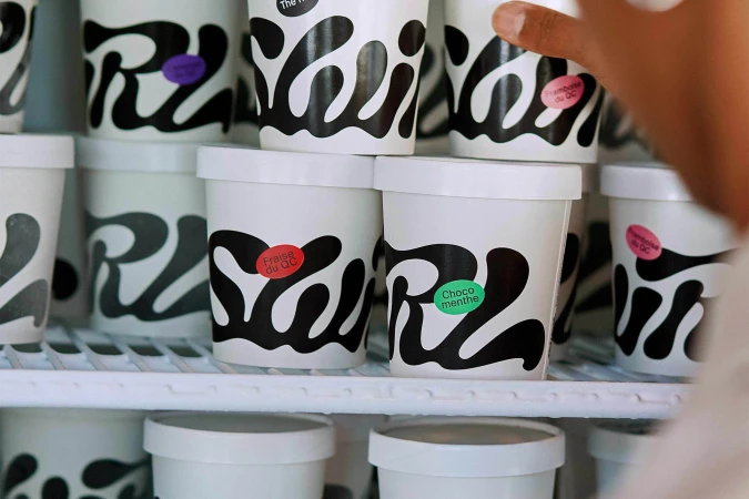 Close-up of white ice cream containers with black designs and colorful flavor labels, arranged on a white rack in a freezer display.