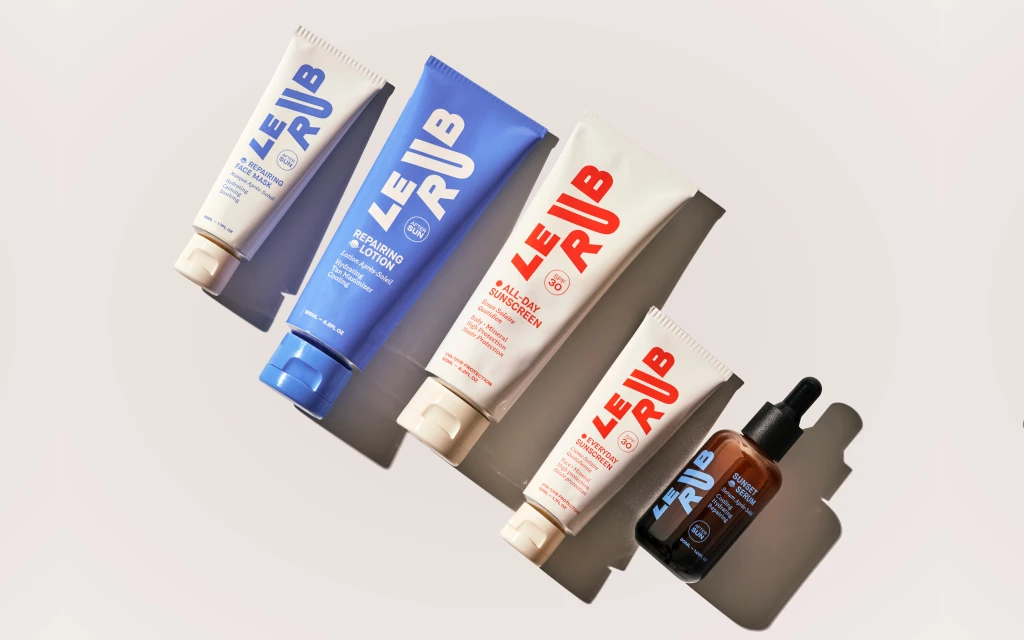 Several tubes and bottles of Le Rub sun and skincare products arranged on a neutral surface in bright, angled studio lighting.