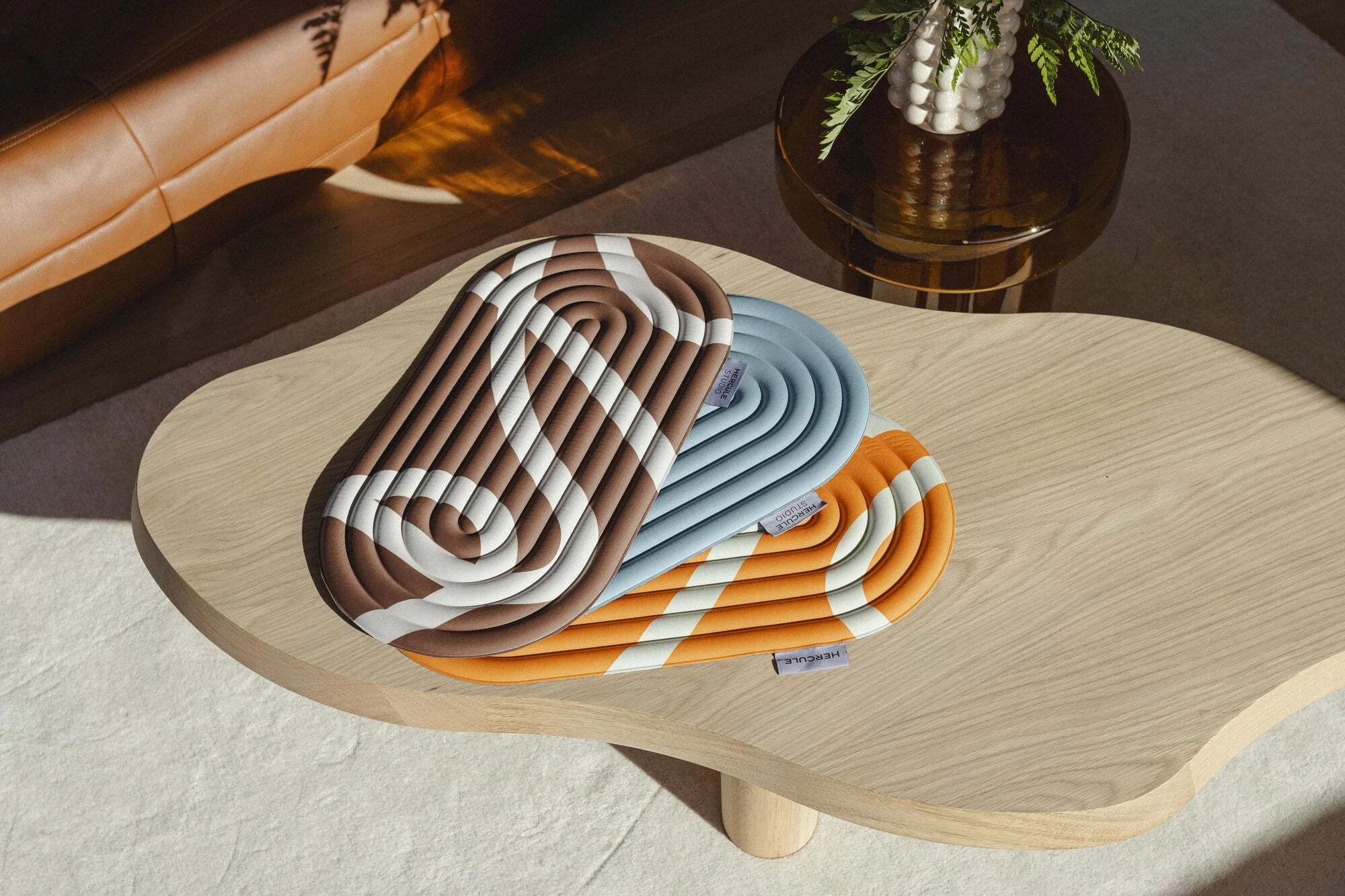 Three patterned trivets in brown, blue, and orange sit atop a light wood coffee table near a leather sofa and a round table with a vase.