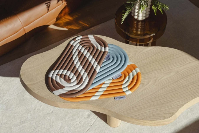 Three patterned trivets in brown, blue, and orange sit atop a light wood coffee table near a leather sofa and a round table with a vase.