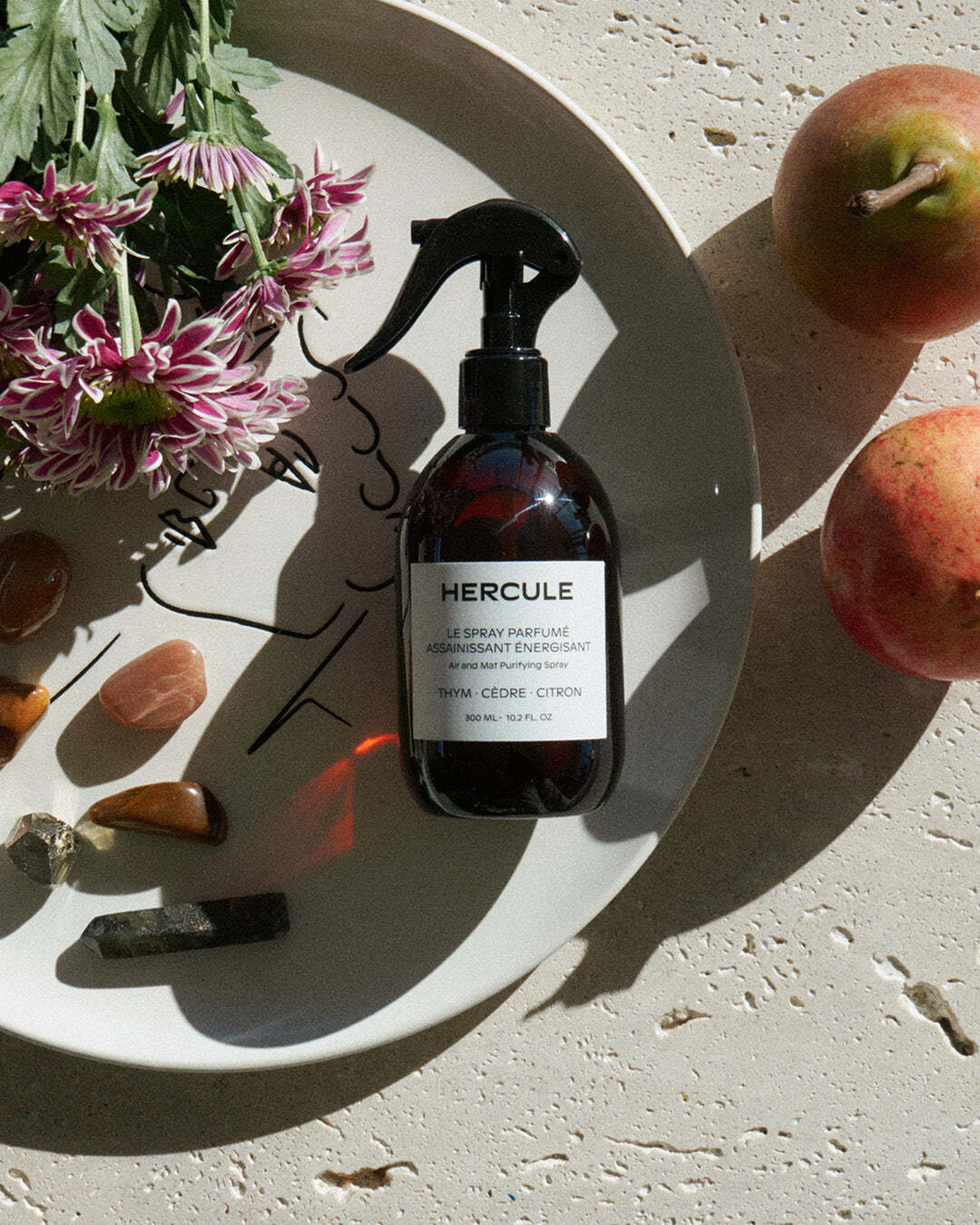A bottle of Herculé Le Spray Parfume sitting on a plate with crystals, apples, and flowers, highlighted by natural sunlight.