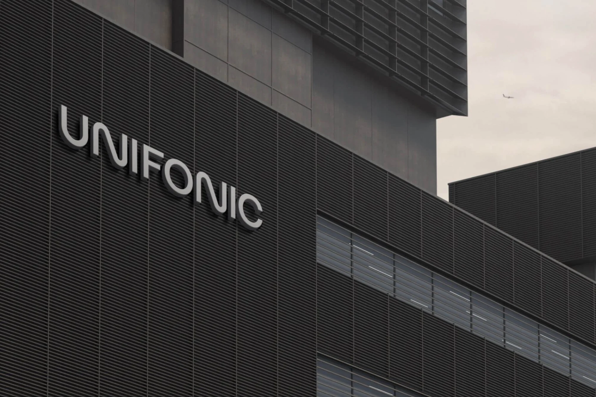 Exterior of a modern building with the sign UNIFONIC. The building has a dark facade and visible windows, with a plane in the sky.
