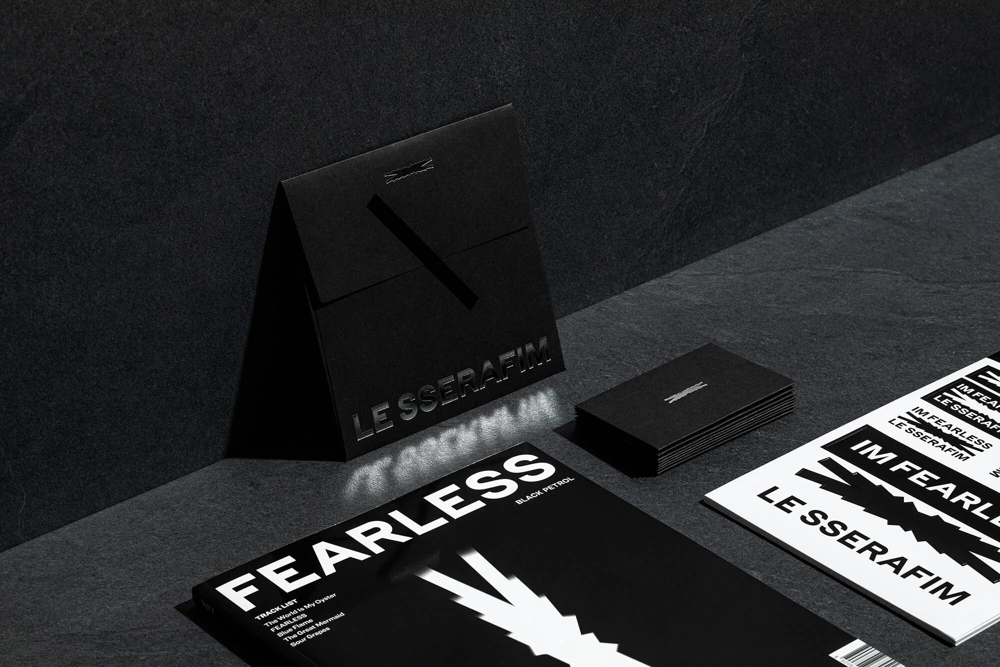 Black and white studio shot featuring LE SSERAFIM magazines, cards, and packaging on a textured dark surface, showcasing design elements.