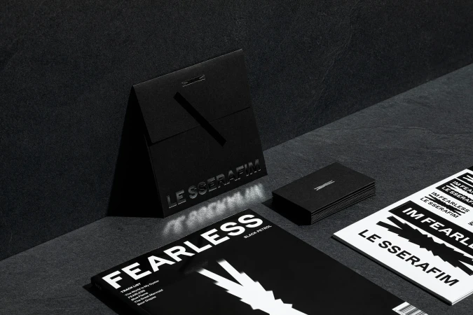 Black and white studio shot featuring LE SSERAFIM magazines, cards, and packaging on a textured dark surface, showcasing design elements.
