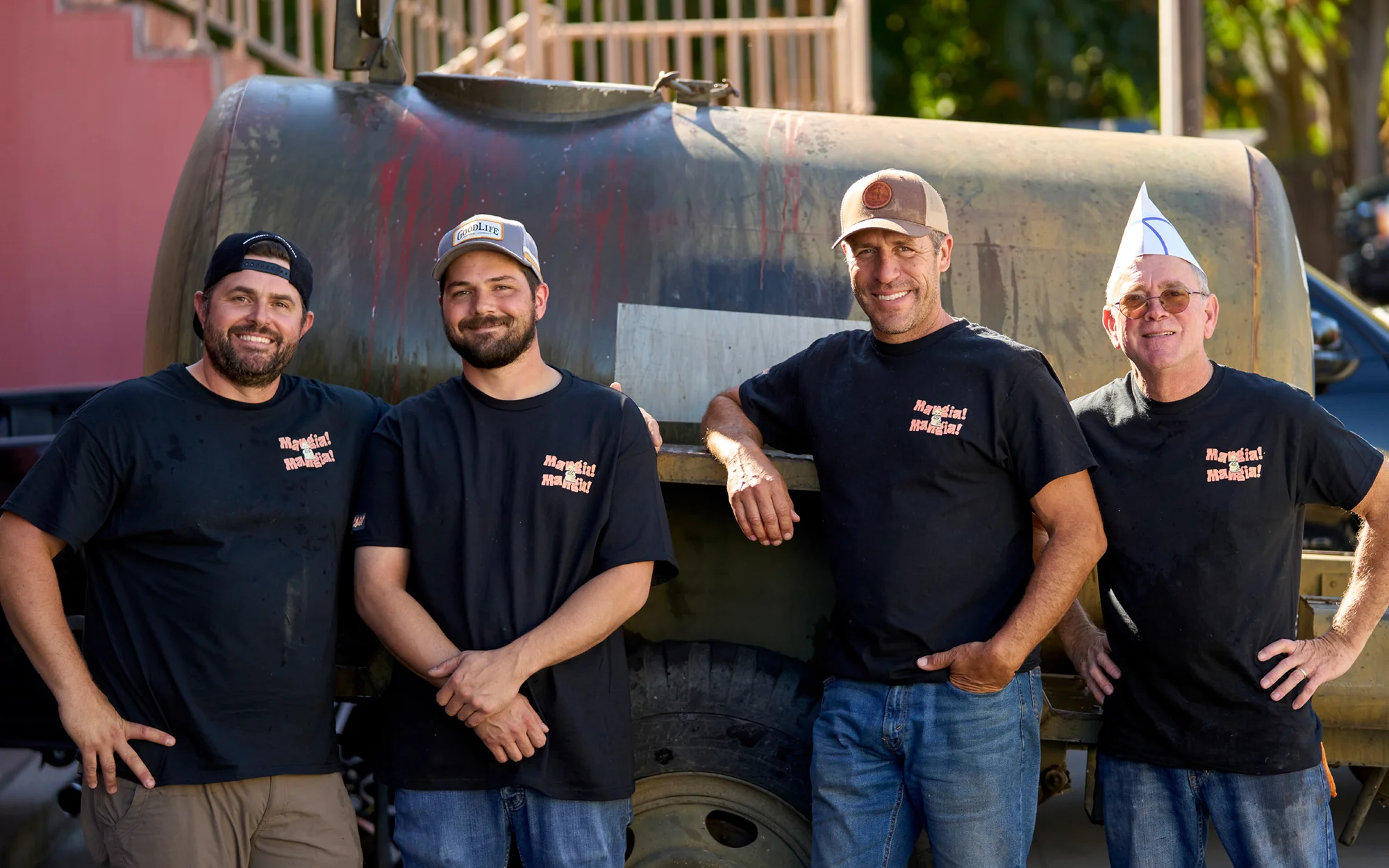Four adult men wearing matching black t-shirts stand in front of an old truck with a large tank in an outdoor setting on a sunny day.
