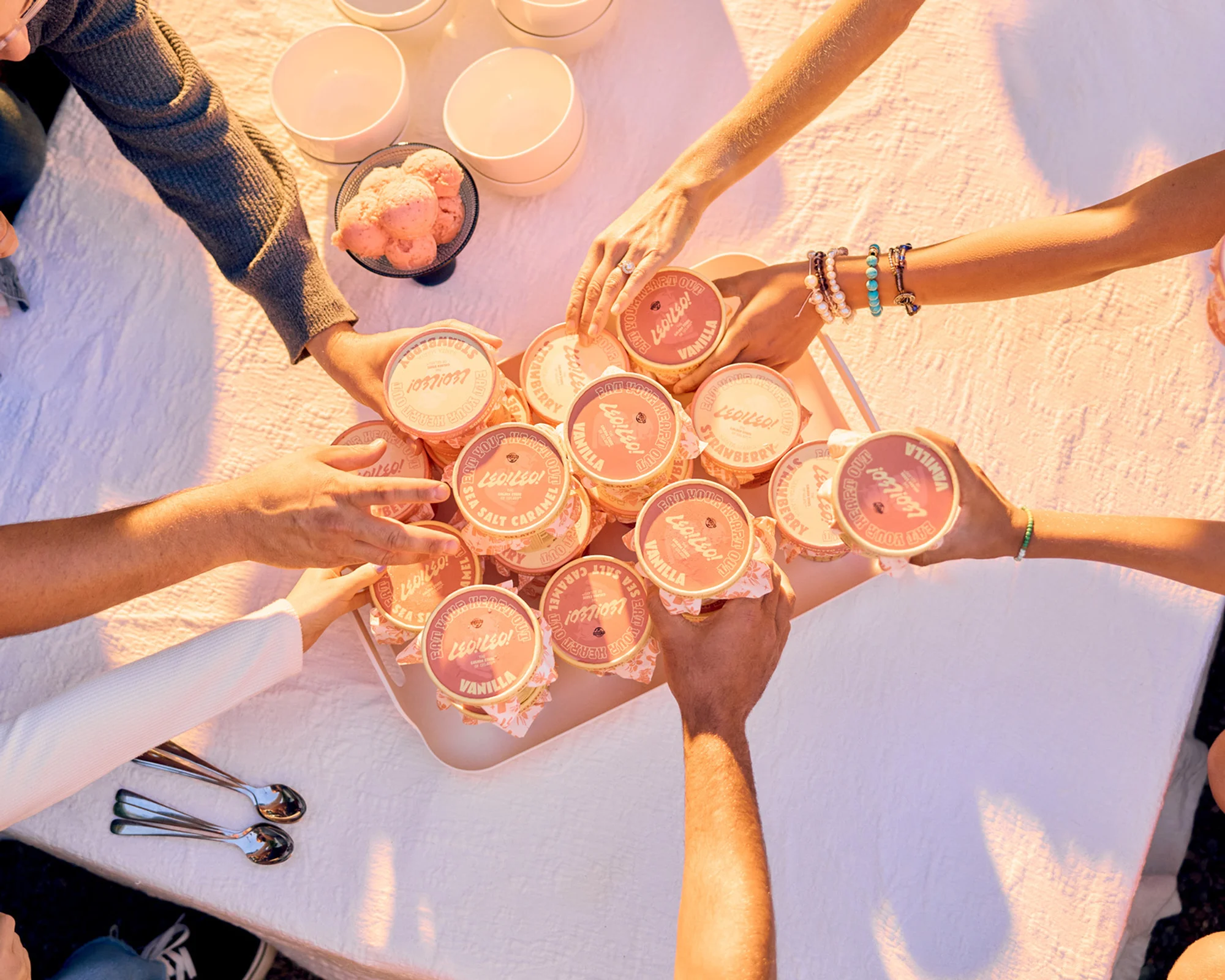 Overhead shot of multiple hands grabbing ice cream containers from a tray on a table, surrounded by small bowls and spoons.