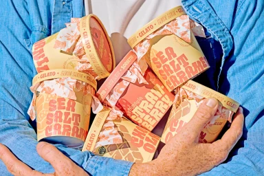 Person wearing a blue denim shirt holding an armful of yellow and red gelato tubs with patterned paper wrap