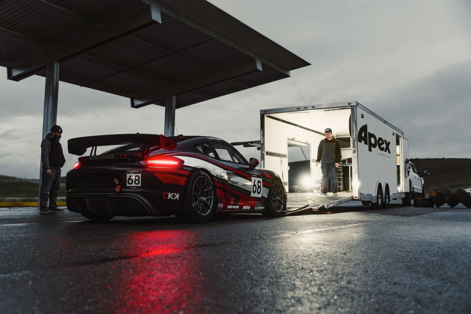 Race car and trailer with two people, outside on wet asphalt, cloudy skies, in a low angle, wide shot on an overcast day.