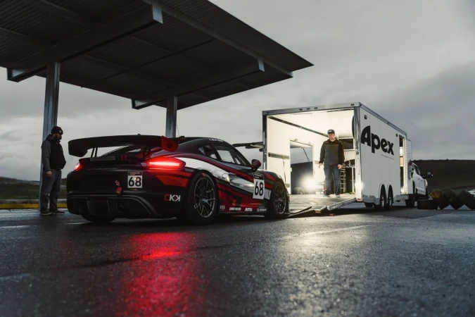 Race car and trailer with two people, outside on wet asphalt, cloudy skies, in a low angle, wide shot on an overcast day.