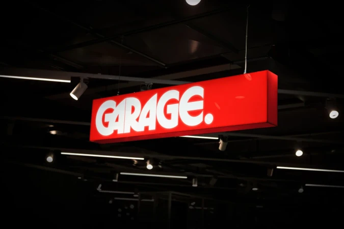 Illuminated red sign reading GARAGE. hanging from the ceiling inside a store, lit by spotlights in a dark environment.