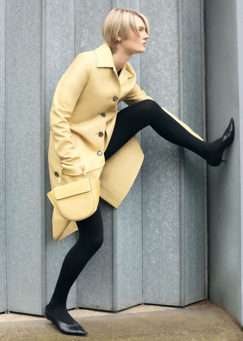 A woman in a yellow trench coat and black tights poses with one leg up against a grey rolling metal door, holding a yellow purse.