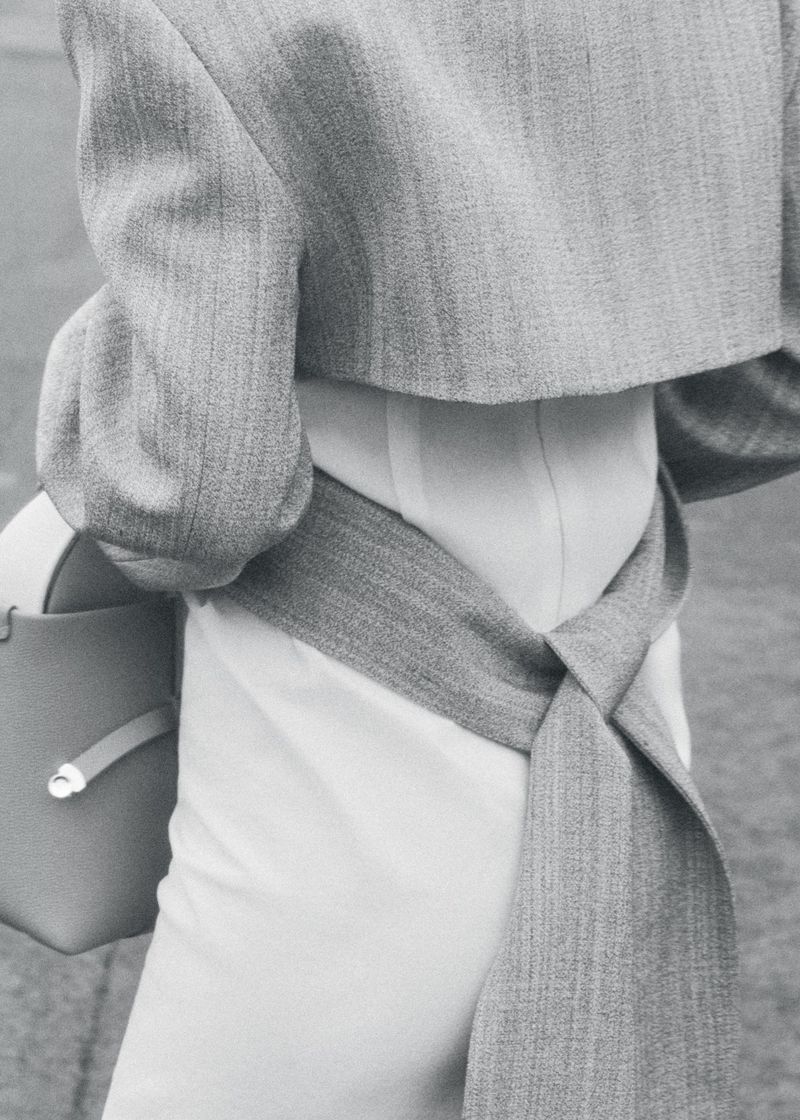 Monochromatic fashion shot of a torso in a white outfit with a light grey jacket, accented by a matching grey sash and handbag.